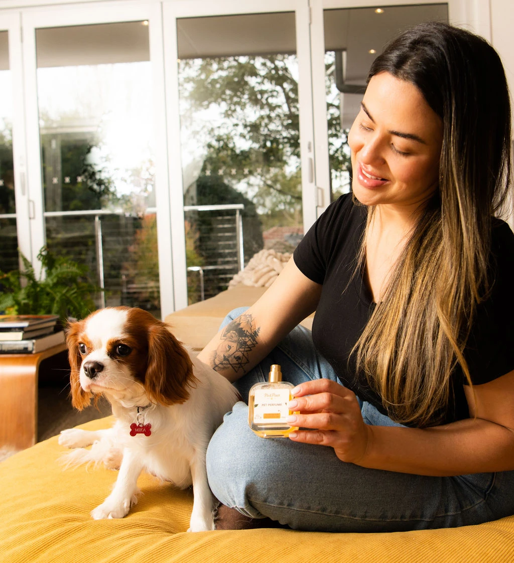 Golden-hour photo of a person sitting with a Cavalier King Charles Spaniel beside them, holding the Pa & Paws Pet Perfume bottle.
